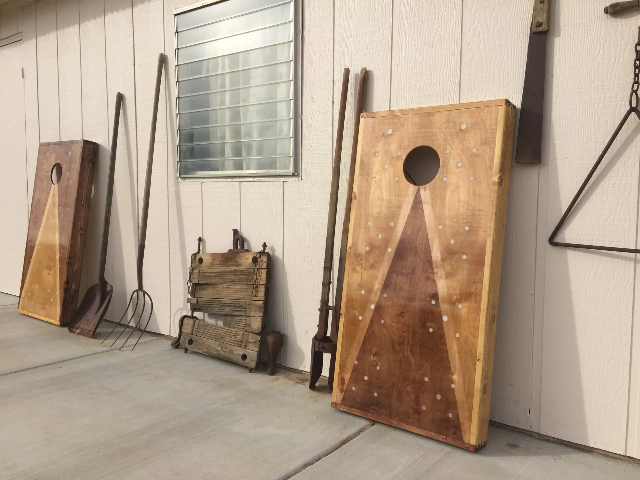 Two rustic YoreBoard cornhole boards, one on each side, leaning against a white building, with a shovel, pitchfork, and other antique farm tools in between them.