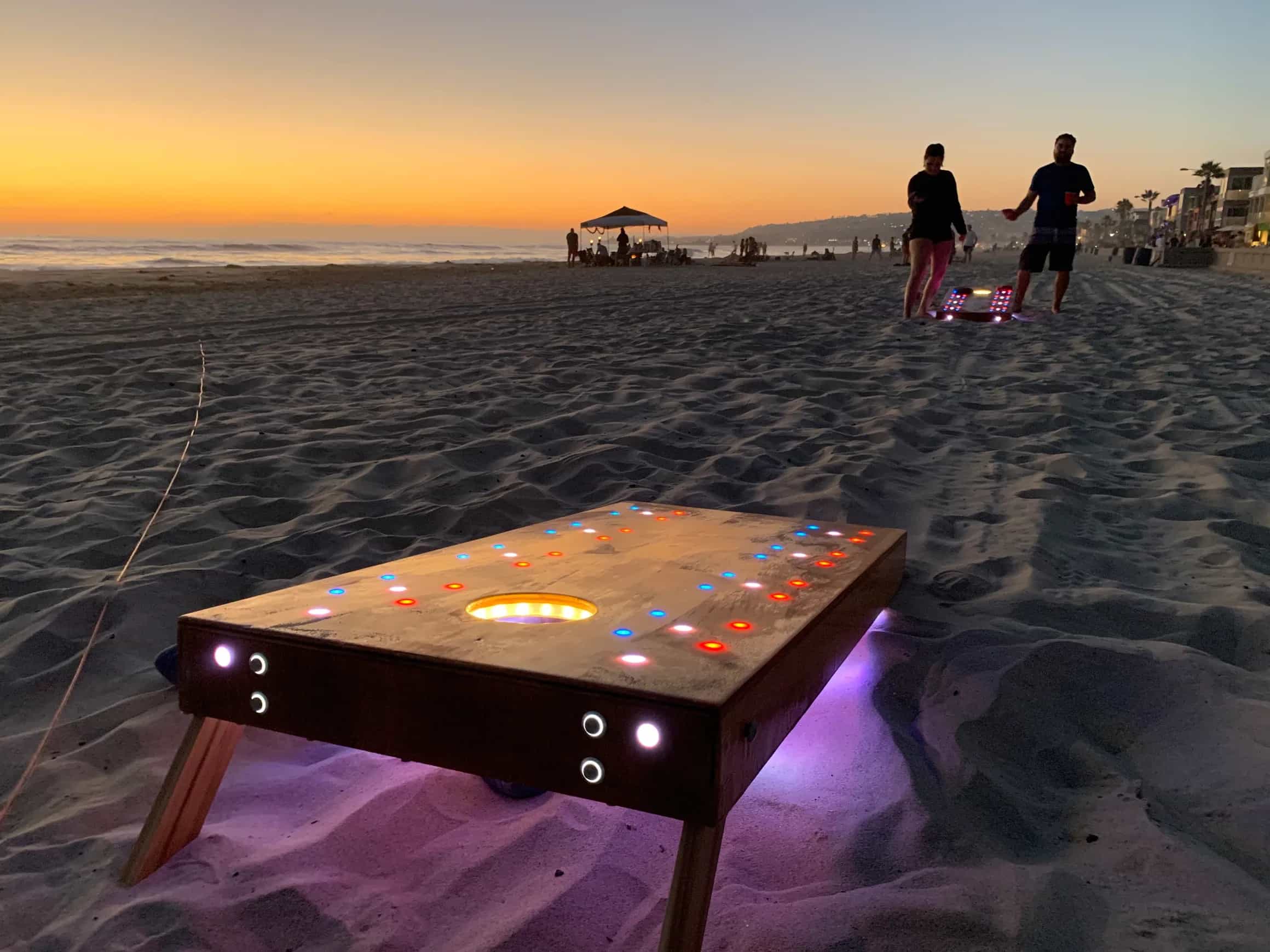 YoreBoard cornhole game in progress on a sandy beach at sunset, with the board illuminated by multi-colored Scoring LEDs and under-board lighting.
