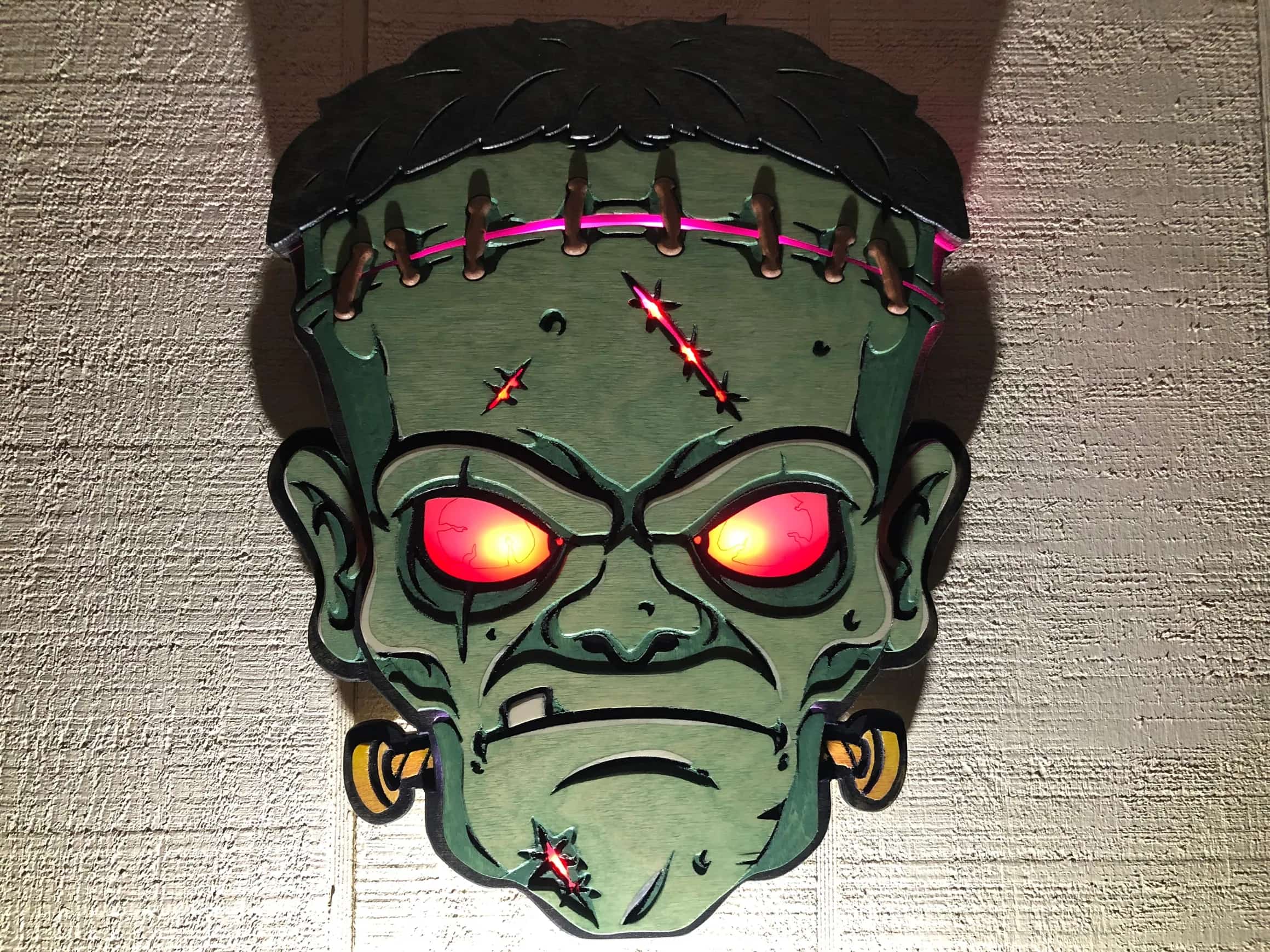 Handcrafted Halloween decoration: a dimensional wooden Frankenstein head with eerie red glowing eyes and pink lights highlighting the stitches on its forehead, showcasing detailed craftsmanship.