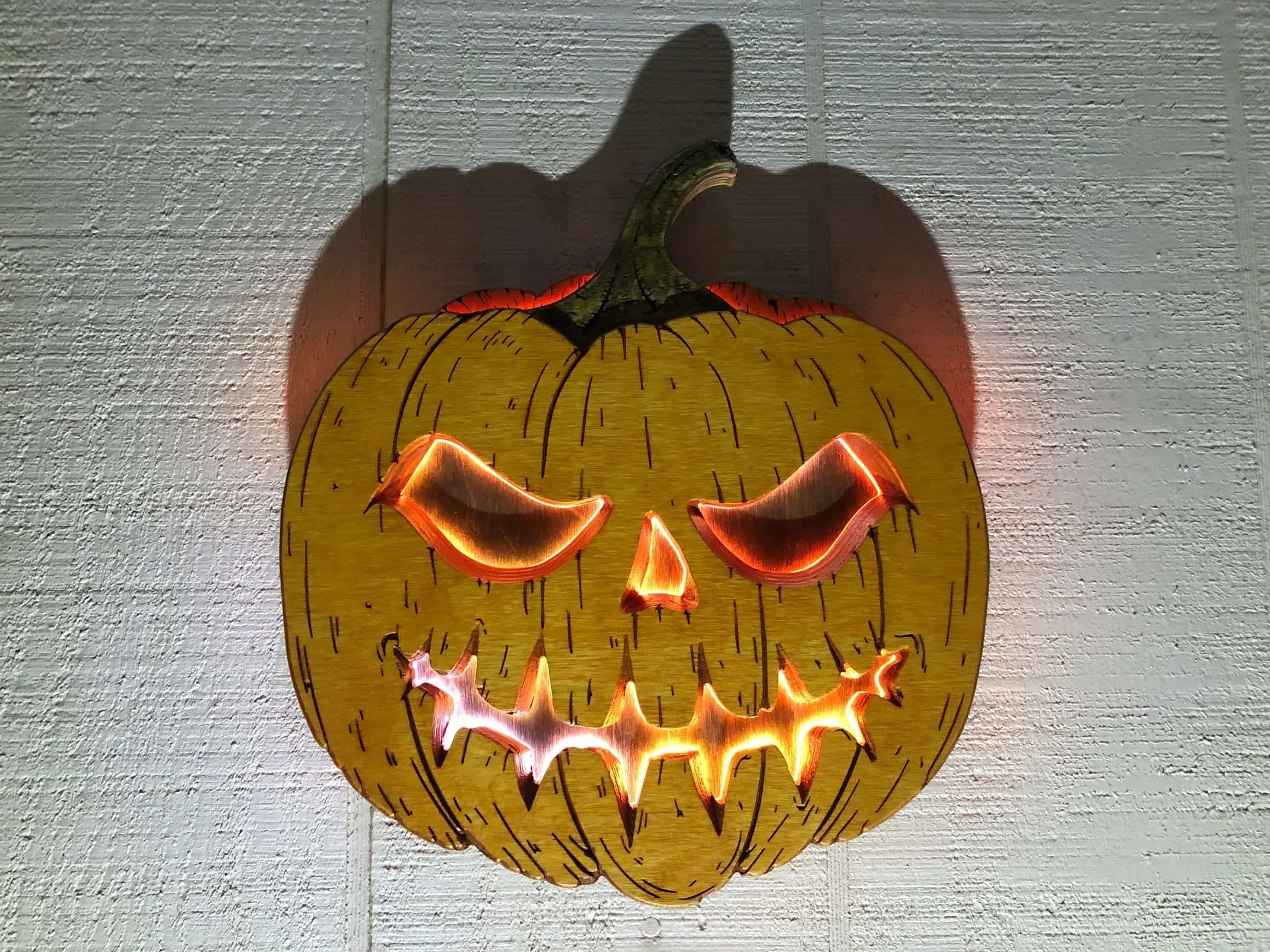 Handcrafted Halloween decoration: a dimensional wooden jackolantern carving with illuminated eyes and a jagged mouth, casting a warm orange glow against a wall.