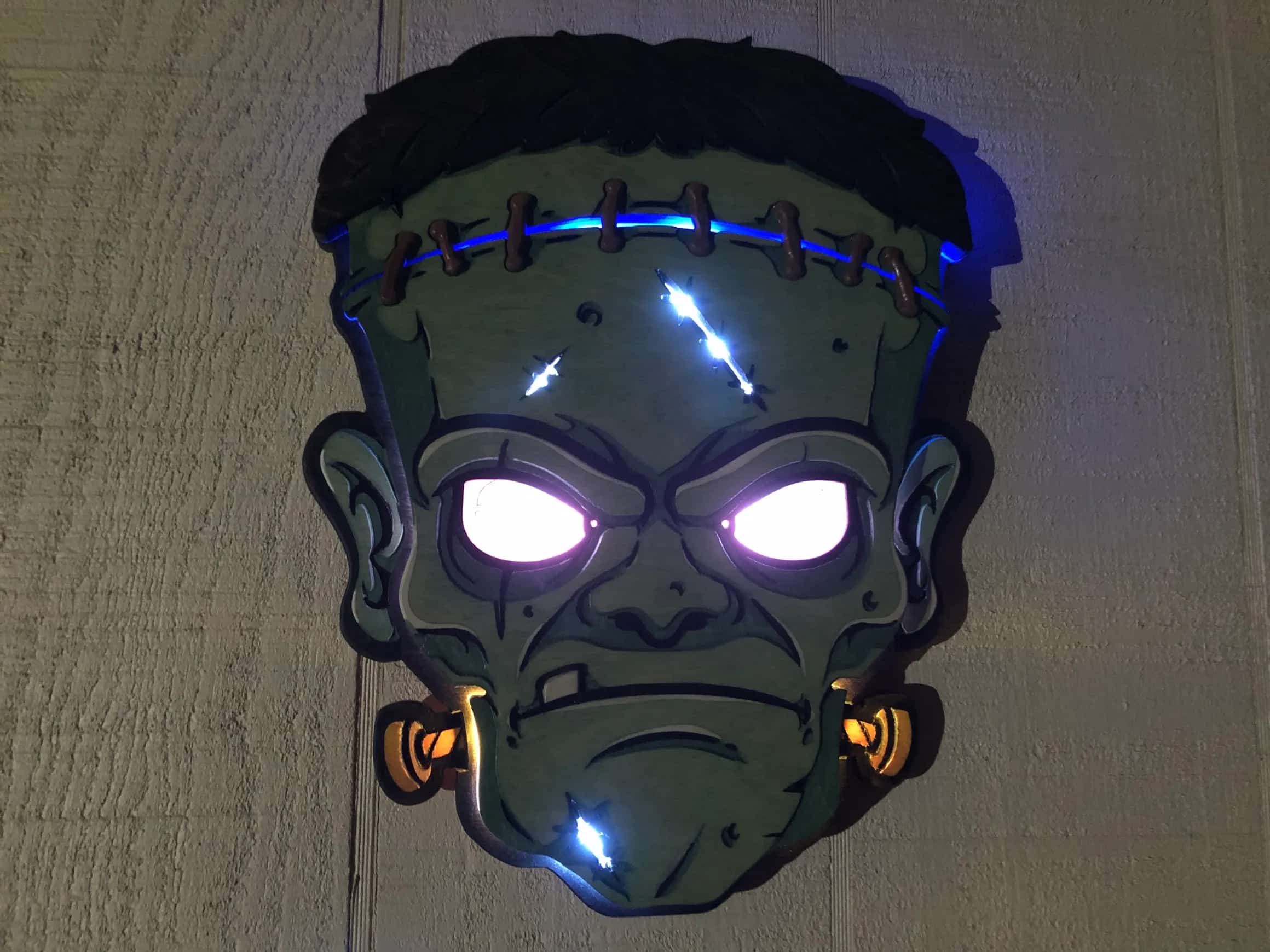 Handcrafted Halloween decoration: a dimensional wooden Frankenstein head with piercing white glowing eyes and blue lights highlighting the stitches on its forehead, showcasing detailed craftsmanship and spooky illumination.