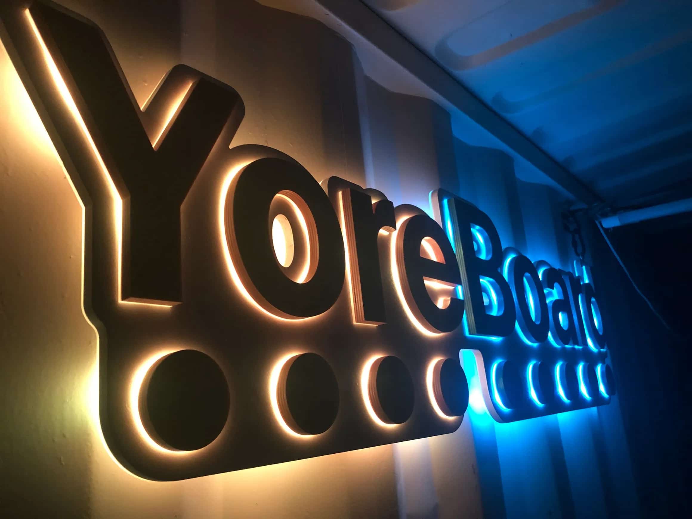 Handcrafted YoreBoard brand sign: a multi-layered wooden display with the company name and circular accents, brilliantly lit by contrasting warm yellow and cool blue LED lights, creating a dynamic visual effect.