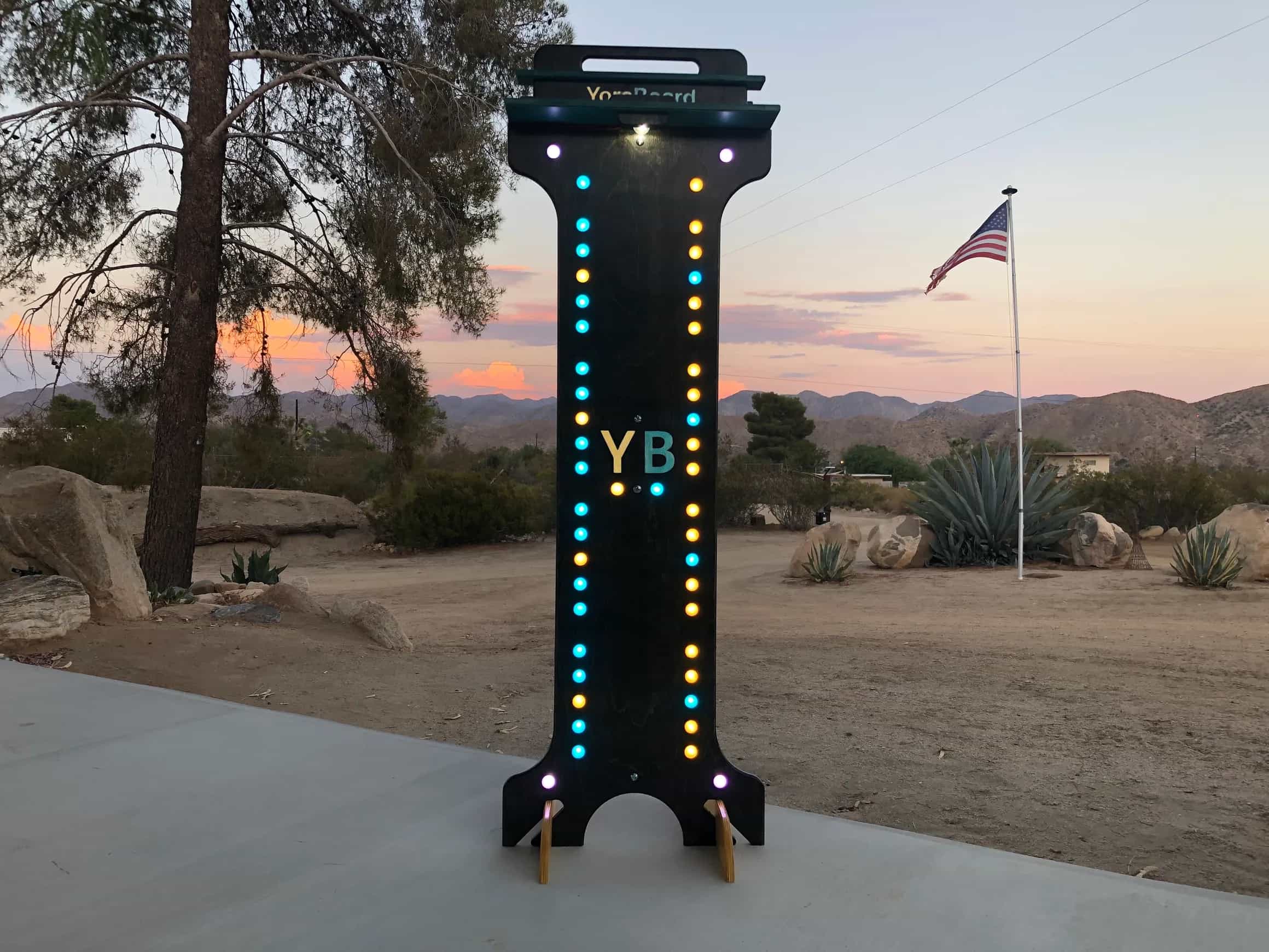 Brightly lit YoreBoard score tower with eye-catching yellow and blue Scoring LEDs and prominent 'YB' brand logo against a twilight sky.