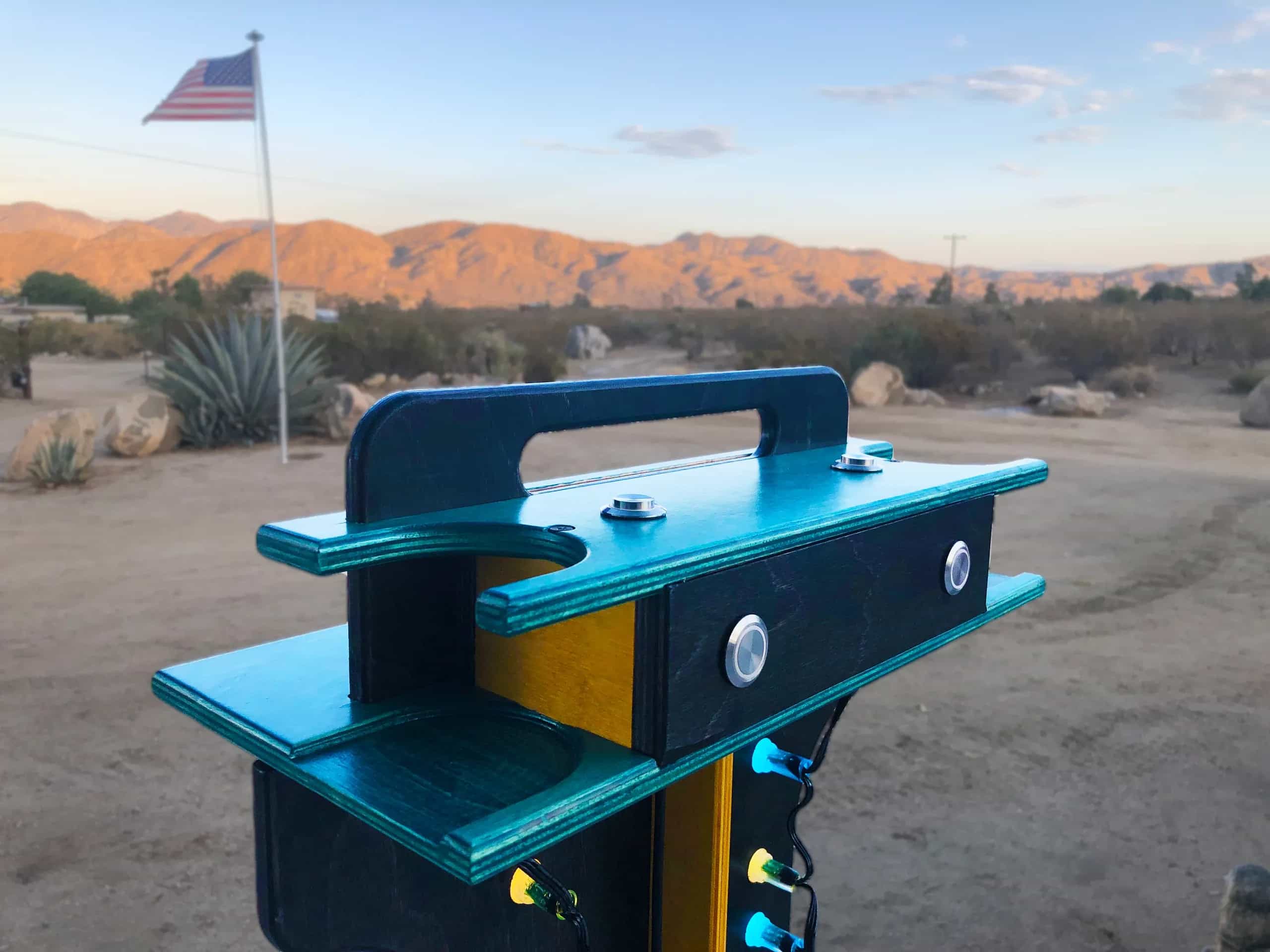 Top section of YoreBoard score tower with integrated carry handle and prominent silver control buttons for score input, set against an outdoor backdrop with an American flag.